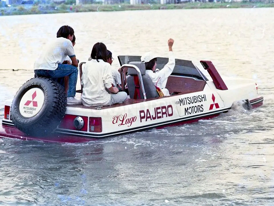 Pandangan dari arah belakang Mitsubishi Pajero El Lago ketika sedang belayar di atas air, mempelihatkan penumpang duduk di atas dek kayu belakang bersebelahan tayar simpanan.